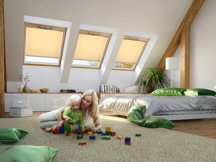 Eine Mutter spielt mit ihrem Kind auf dem Boden des Schlafzimmers, im Hintergrund 3 Dachfenster aus Holz mit fast geschlossenen Rollos innen
